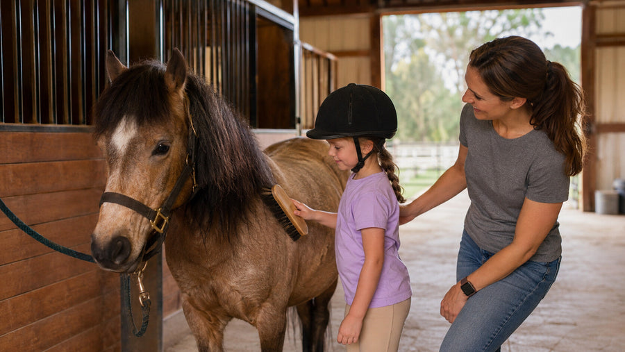 Young helmeted rider learning to groom a pony with adult supervision